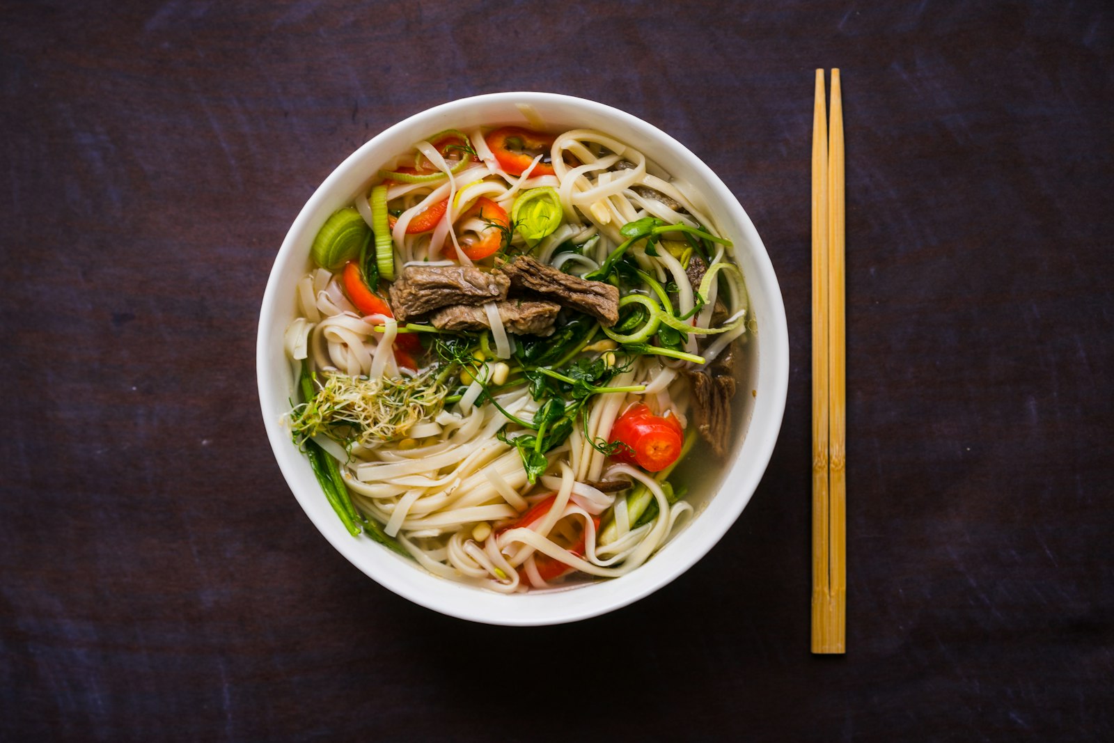 Overhead view of a bowl of beef pho with herbs, chili and chopsticks