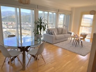 A modern living room with a large glass table surrounded by four chairs. The room features hardwood floors, a cozy gray sofa with striped cushions, and a pair of nested coffee tables on a textured rug. Large windows offer a panoramic view of a cityscape and mountains, while a potted plant adds a touch of greenery.