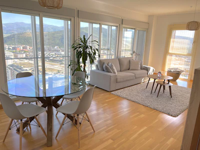 A modern living room with a large glass table surrounded by four chairs. The room features hardwood floors, a cozy gray sofa with striped cushions, and a pair of nested coffee tables on a textured rug. Large windows offer a panoramic view of a cityscape and mountains, while a potted plant adds a touch of greenery.