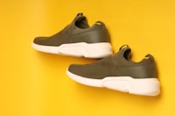 Two olive green sneakers with white soles are positioned in a diagonal layout on a vibrant yellow background. The sneakers have a streamlined design with mesh detailing on the upper part and a loop at the back for easy wear.
