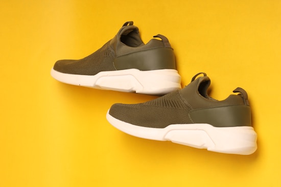 Two olive green sneakers with white soles are positioned in a diagonal layout on a vibrant yellow background. The sneakers have a streamlined design with mesh detailing on the upper part and a loop at the back for easy wear.