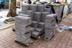 Stacks of gray paving stones are neatly arranged on wooden pallets, surrounded by construction materials such as plastic sheeting and other supplies. The setting appears to be an outdoor construction site with a tree visible and parts of unfinished pavement around.