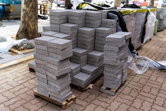 Stacks of gray paving stones are neatly arranged on wooden pallets, surrounded by construction materials such as plastic sheeting and other supplies. The setting appears to be an outdoor construction site with a tree visible and parts of unfinished pavement around.