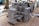 Stacks of gray paving stones are neatly arranged on wooden pallets, surrounded by construction materials such as plastic sheeting and other supplies. The setting appears to be an outdoor construction site with a tree visible and parts of unfinished pavement around.