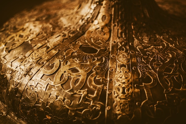 Intricate brass or bronze metal surface with detailed engravings and patterns. The design features various abstract shapes and symbols, possibly of historical or cultural significance. The surface appears aged with a warm, golden hue, and has a reflective quality.