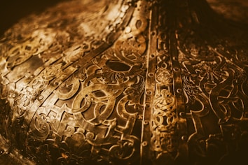 Intricate brass or bronze metal surface with detailed engravings and patterns. The design features various abstract shapes and symbols, possibly of historical or cultural significance. The surface appears aged with a warm, golden hue, and has a reflective quality.