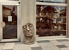 A large stone sculpture of a face is positioned on the ground outside a storefront with glass windows. The store appears to sell gifts and decorative items, as indicated by reflections and signs. The facade includes a visible door, concrete walls, and a tiled pathway leading to the entrance.