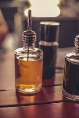 A close-up of two glass bottles on a wooden surface, one filled with an amber liquid and the other dark, possibly containing oil and vinegar. The bottles have metal spouts, and a blurred background indicates a warm ambient setting.