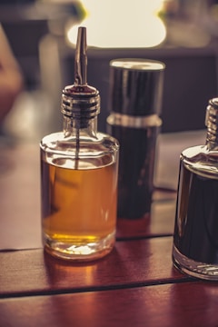 A close-up of two glass bottles on a wooden surface, one filled with an amber liquid and the other dark, possibly containing oil and vinegar. The bottles have metal spouts, and a blurred background indicates a warm ambient setting.