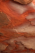A close-up view of layered rock formations showcasing intricate patterns and textures. The rock has swirling bands and striations in hues of red, orange, and brown, likely due to the natural erosion and sediment deposition over time.