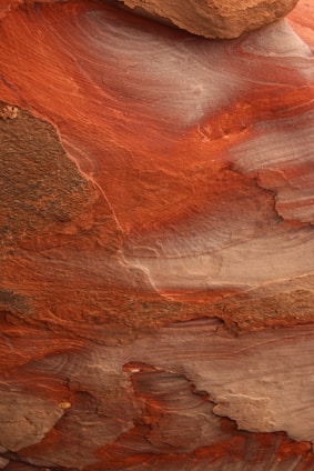 A close-up view of layered rock formations showcasing intricate patterns and textures. The rock has swirling bands and striations in hues of red, orange, and brown, likely due to the natural erosion and sediment deposition over time.