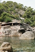 A rustic building with a wooden roof is nestled on a rocky hillside covered in lush green trees. Below the building, people are sunbathing on rocks near the water. The foreground showcases clear water with visible rock formations both in and out of the water.