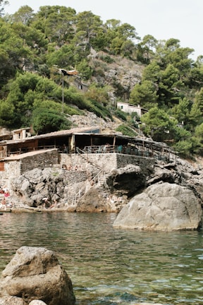 A rustic building with a wooden roof is nestled on a rocky hillside covered in lush green trees. Below the building, people are sunbathing on rocks near the water. The foreground showcases clear water with visible rock formations both in and out of the water.