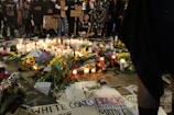 An assembly of people stands solemnly around a memorial on the ground. The memorial comprises numerous lit candles, flowers, and various signs bearing messages of solidarity and protest. Many of the signs display phrases such as 'Black Lives Matter' and 'Black Trans Lives Matter'. The scene is illuminated by the soft glow of candlelight, creating a poignant atmosphere.
