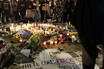 An assembly of people stands solemnly around a memorial on the ground. The memorial comprises numerous lit candles, flowers, and various signs bearing messages of solidarity and protest. Many of the signs display phrases such as 'Black Lives Matter' and 'Black Trans Lives Matter'. The scene is illuminated by the soft glow of candlelight, creating a poignant atmosphere.