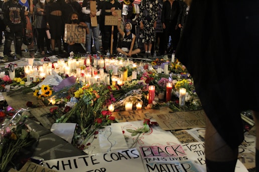 An assembly of people stands solemnly around a memorial on the ground. The memorial comprises numerous lit candles, flowers, and various signs bearing messages of solidarity and protest. Many of the signs display phrases such as 'Black Lives Matter' and 'Black Trans Lives Matter'. The scene is illuminated by the soft glow of candlelight, creating a poignant atmosphere.