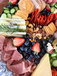 A charcuterie board adorned with a variety of foods, including slices of cheese, cured meats, crackers, strawberries, blueberries, cucumber slices, nuts, a small bowl of creamy dip, and sprigs of rosemary for garnish.