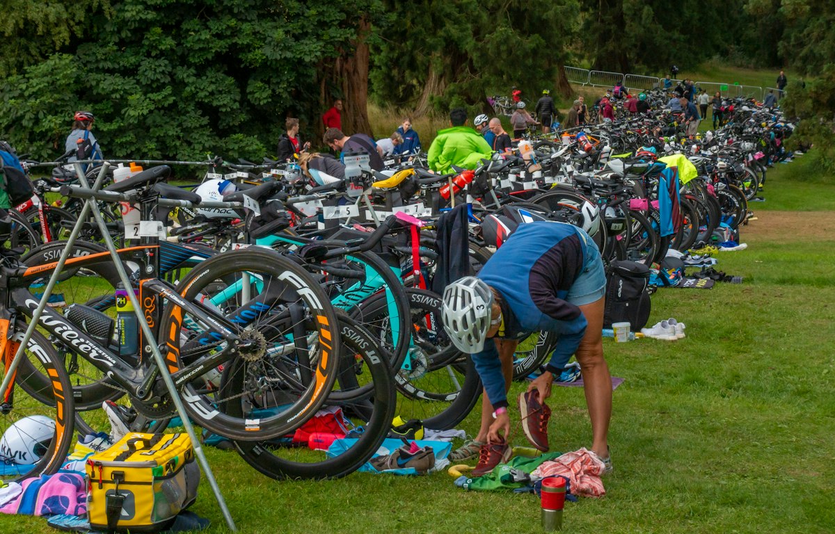 Parc à vélos de triathlon sur l'herbe, vélos alignés sur les racks et athlète qui prépare son matériel