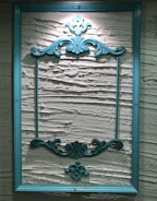 An ornate, teal-colored wooden frame hangs on a textured, cream-colored wall. The frame features intricate, decorative carvings at the top and bottom, adding a classic and elegant touch to the overall appearance.