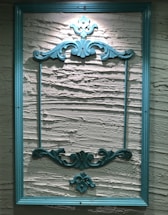 An ornate, teal-colored wooden frame hangs on a textured, cream-colored wall. The frame features intricate, decorative carvings at the top and bottom, adding a classic and elegant touch to the overall appearance.