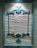 An ornate, teal-colored wooden frame hangs on a textured, cream-colored wall. The frame features intricate, decorative carvings at the top and bottom, adding a classic and elegant touch to the overall appearance.