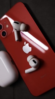 A red smartphone with an Apple logo on the back lies on a dark surface. Two wireless earbuds rest on the phone, and a charging case is nearby.