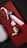 A red smartphone with an Apple logo on the back lies on a dark surface. Two wireless earbuds rest on the phone, and a charging case is nearby.