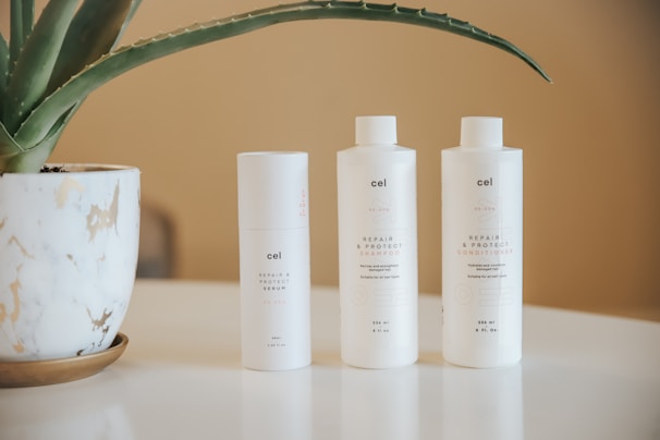 A marble-patterned planter with a green aloe vera plant sits on a table. Next to it are three white bottles labeled as hair care products: serum, shampoo, and conditioner. The background is a soft beige color.