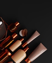 A collection of various cosmetic containers and bottles arranged neatly against a dark background. The colors of the containers range from muted beige to shiny bronze, featuring sleek and modern designs.