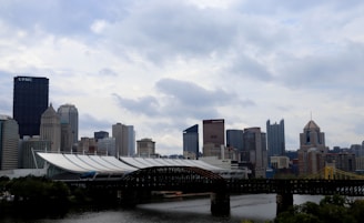 A cityscape featuring a variety of modern and historic buildings in a skyline. The scene includes a large black skyscraper with the letters 'UPMC' on it and other notable high-rise buildings. An iron bridge spans across a river in the foreground, with lush greenery along the riverbanks. The sky is overcast with thick cloud cover, giving a dramatic backdrop to the urban setting.