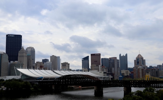 A cityscape featuring a variety of modern and historic buildings in a skyline. The scene includes a large black skyscraper with the letters 'UPMC' on it and other notable high-rise buildings. An iron bridge spans across a river in the foreground, with lush greenery along the riverbanks. The sky is overcast with thick cloud cover, giving a dramatic backdrop to the urban setting.