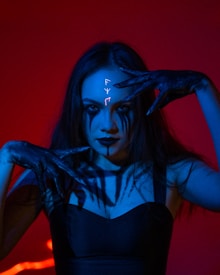 A woman with long dark hair and striking makeup features prominently against a vibrant red background. Her hands are positioned around her face, accentuating her black lipstick and dark eye makeup. Mysterious symbolic markings appear on her forehead, glowing with an ethereal light. The image is dramatically lit with a blue hue, creating a striking contrast with the red backdrop.