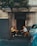 Two elderly men are sitting on folding chairs in front of a building, engaged in conversation. One man gestures with his hand while holding a cigarette, and the other holds some papers. A tree partially obscures the view of the men, and a parked car is visible on the right side of the image. The building has an aged appearance with faded signage above.