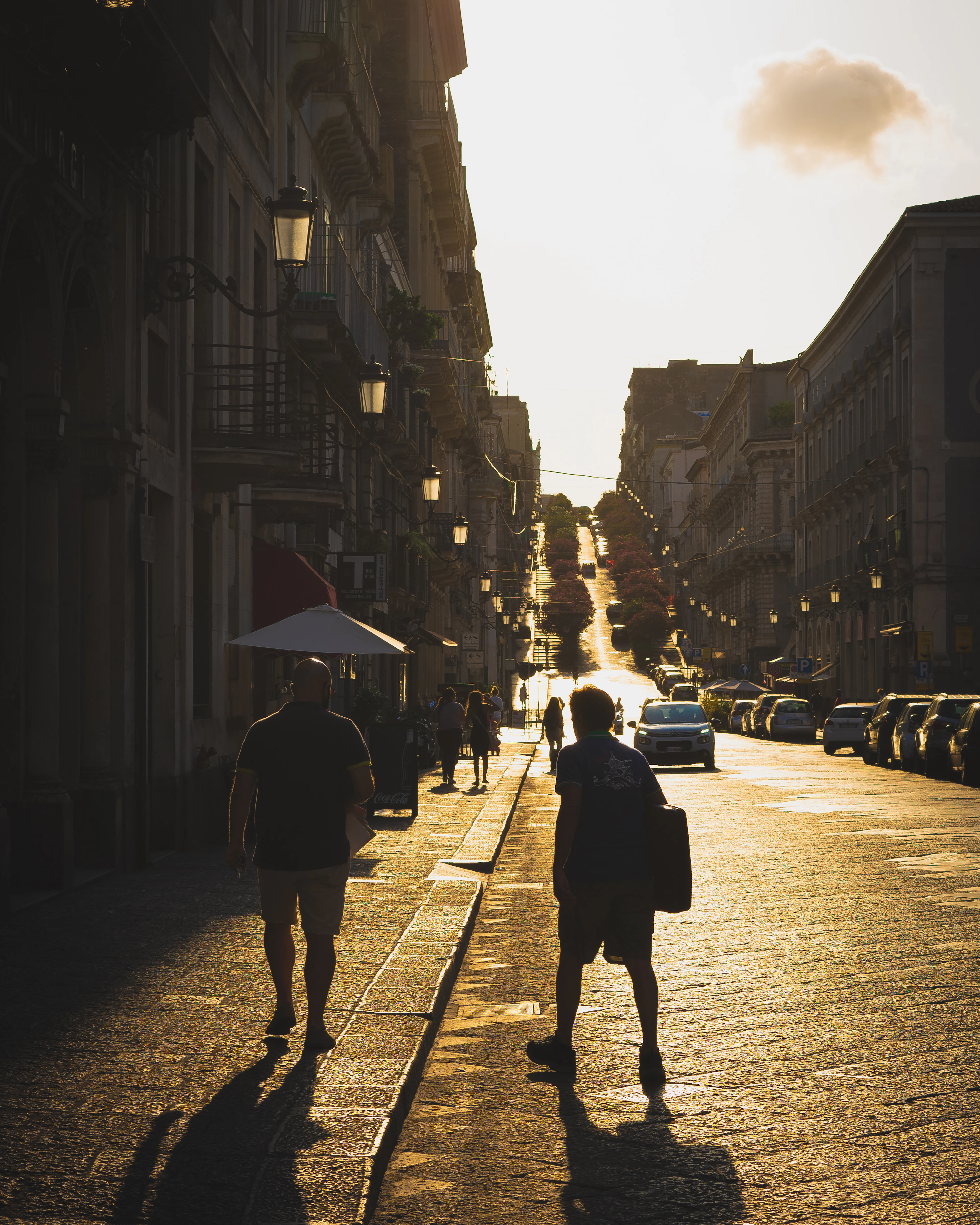 Pedestrians are silhouetted in golden evening light on a long Catania avenue.