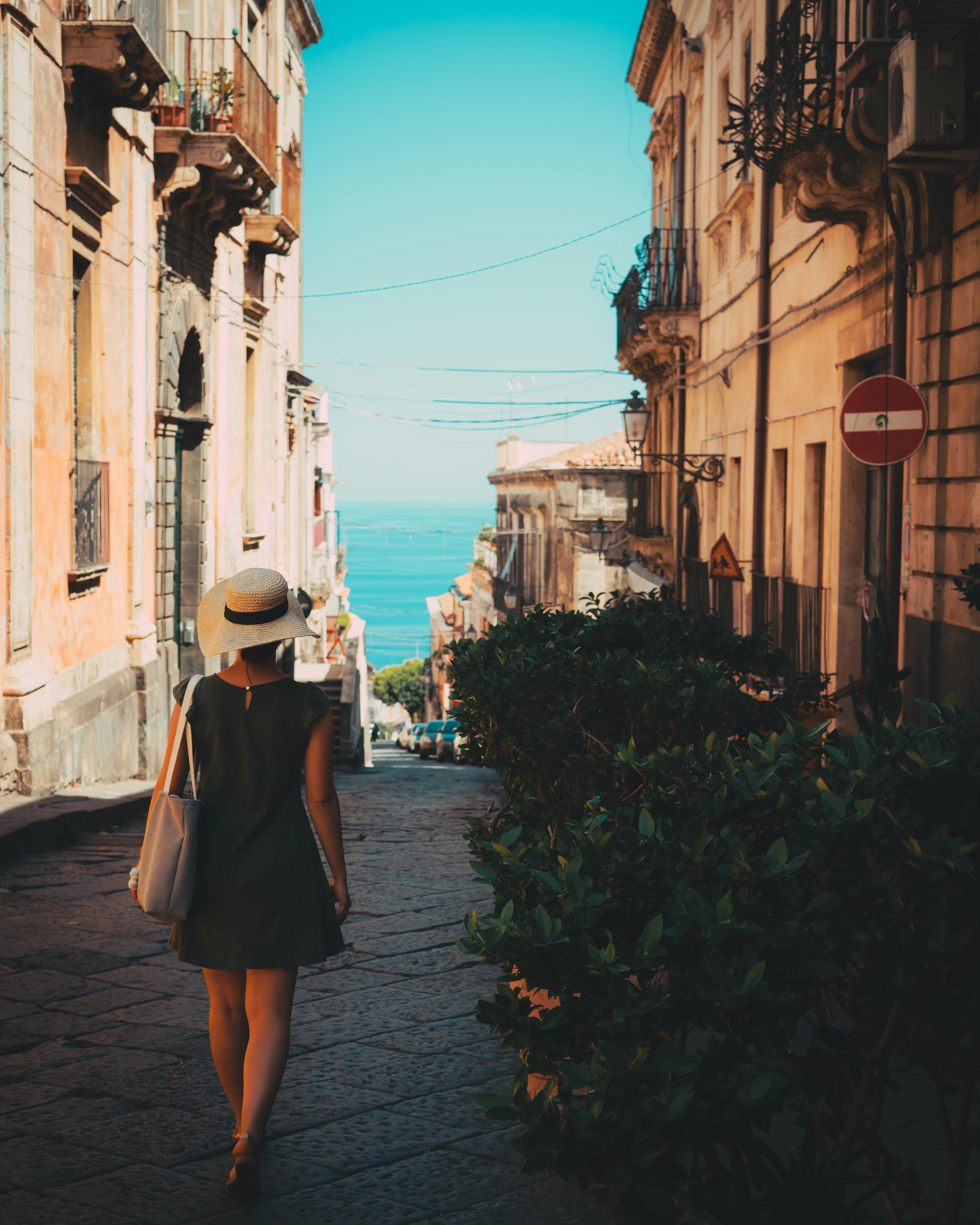 A woman in a straw hat walks down a Catania street that opens onto a view of the sea.