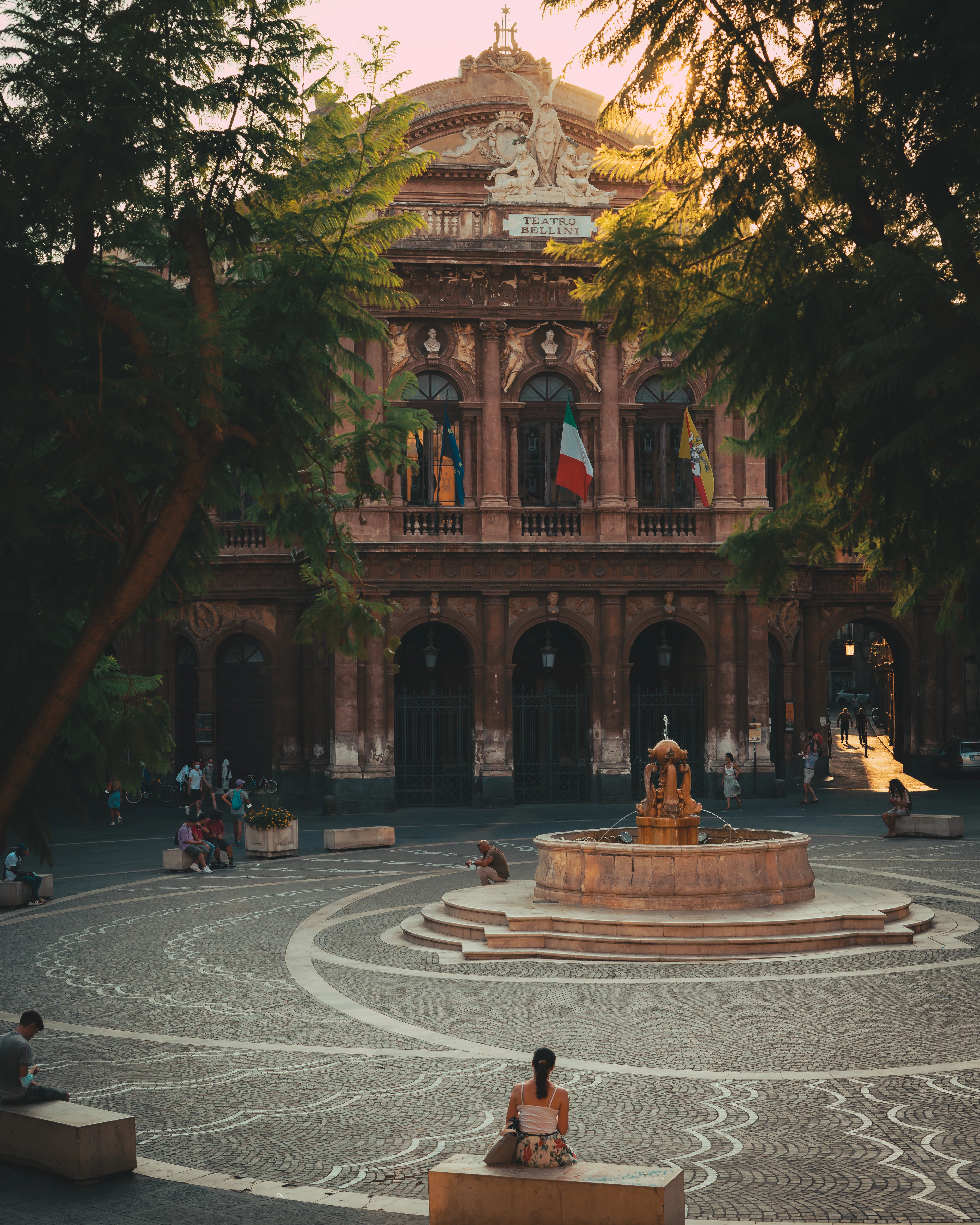 The ornate facade of Catania's Teatro Massimo Bellini above a stone fountain and swirling paved square, framed by trees at dusk