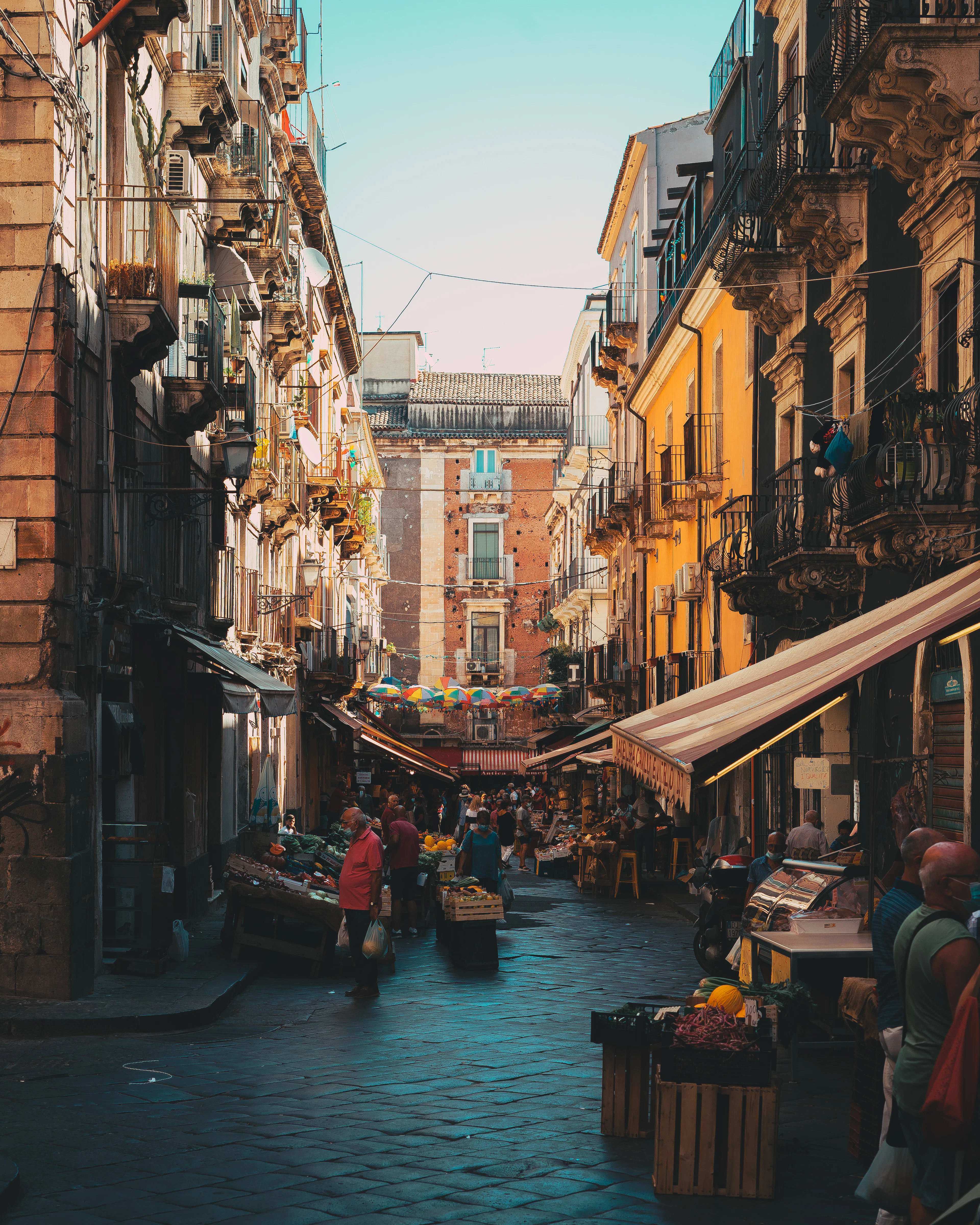 A narrow market street in Catania lined with produce crates, striped awnings and shoppers, tall balconied buildings closing in on both sides