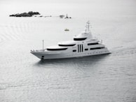 A large white yacht is sailing through calm waters, with small rocky formations and a buoy visible in the background. The sky appears overcast, casting a serene, grey light over the scene.