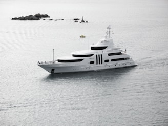 A large white yacht is sailing through calm waters, with small rocky formations and a buoy visible in the background. The sky appears overcast, casting a serene, grey light over the scene.