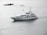 A large white yacht is sailing through calm waters, with small rocky formations and a buoy visible in the background. The sky appears overcast, casting a serene, grey light over the scene.