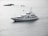 A large white yacht is sailing through calm waters, with small rocky formations and a buoy visible in the background. The sky appears overcast, casting a serene, grey light over the scene.