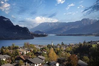 A picturesque lakeside village surrounded by lush greenery and mountains. The village houses have traditional architecture with sloped roofs, and a prominent church tower rises among them. In the background, a serene lake reflects the blue sky and is bordered by towering mountains.