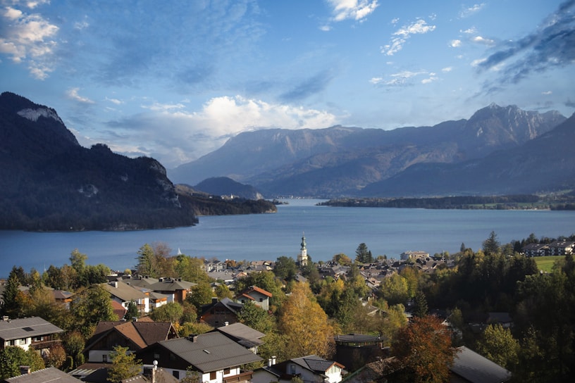 A picturesque lakeside village surrounded by lush greenery and mountains. The village houses have traditional architecture with sloped roofs, and a prominent church tower rises among them. In the background, a serene lake reflects the blue sky and is bordered by towering mountains.