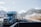 A blue semi-truck is driving on a highway with a mountainous backdrop covered with a light dusting of snow. Several other vehicles are visible on the road, including another truck and a car. There are clear blue skies with some scattered clouds.