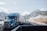 A blue semi-truck is driving on a highway with a mountainous backdrop covered with a light dusting of snow. Several other vehicles are visible on the road, including another truck and a car. There are clear blue skies with some scattered clouds.