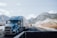 A blue semi-truck is driving on a highway with a mountainous backdrop covered with a light dusting of snow. Several other vehicles are visible on the road, including another truck and a car. There are clear blue skies with some scattered clouds.