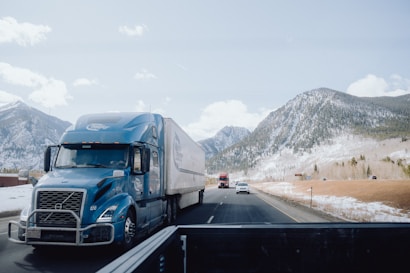 A blue semi-truck is driving on a highway with a mountainous backdrop covered with a light dusting of snow. Several other vehicles are visible on the road, including another truck and a car. There are clear blue skies with some scattered clouds.