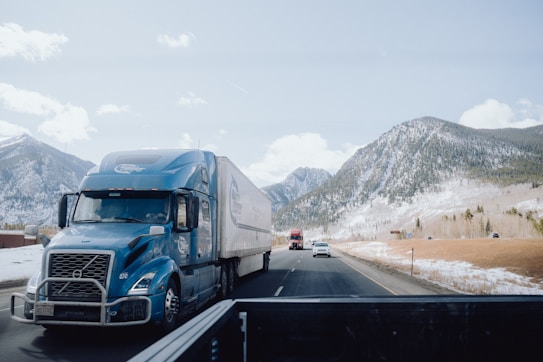 A blue semi-truck is driving on a highway with a mountainous backdrop covered with a light dusting of snow. Several other vehicles are visible on the road, including another truck and a car. There are clear blue skies with some scattered clouds.