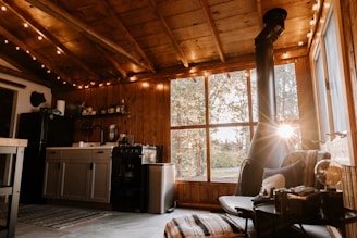 A cozy wooden cabin interior with warm lighting and rustic decor. The room features a small kitchen area with a sink, stove, and refrigerator, along with open shelving holding various kitchen items. A large window allows natural light to pour in, highlighting the wooden walls and ceiling. A modern chair and small table sit near a freestanding stove, giving the space a comfortable and inviting atmosphere.