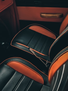 The image shows the interior of a car, focusing on the seats that have a sleek design. They are upholstered in a combination of black and orange leather with decorative stripes. The side panel of the door is also visible, featuring a wooden handle.